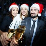 1 Male and 2 Female Young Professionals Wearing Santa Hats Toasting With Champaign In A Photo Booth At A Corporate Holiday Party With Live Music