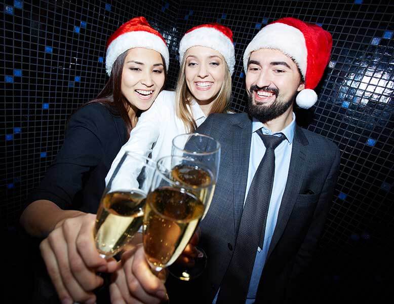 1 Male and 2 Female Young Professionals Wearing Santa Hats Toasting With Champaign In A Photo Booth At A Corporate Holiday Party With Live Music